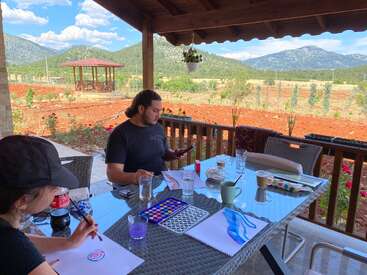 Two people sit at a patio table outdoors, painting with watercolors. Mountains and a gazebo are in the background, beneath a bright blue sky with clouds.