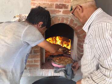 Two people are baking in a brick, wood-fired oven. One is placing or removing a dish inside, while flames burn brightly in the background.