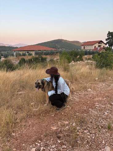 A person wearing a wide-brimmed hat kneels down, hugging a large brown dog in a grassy field, with houses and hills in the background.