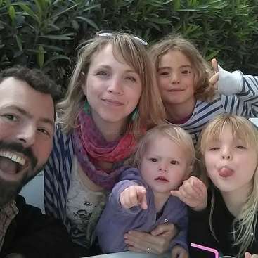 Das Bild zeigt eine fünfköpfige Familie mit zwei Erwachsenen und drei Kindern, die für ein Selfie vor einer grünen Hecke posiert, mit einem Wasserzeichen in der Mitte.