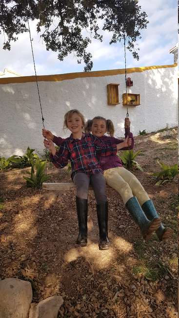 The image depicts two young girls sitting on a wooden swing, holding hands, and smiling at the camera, in a yard with a white wall and a tree.