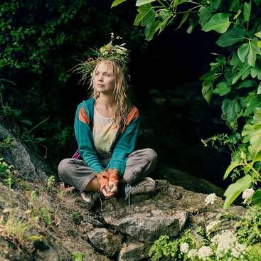 A woman sits cross-legged on a rock by a cave, wearing a crown of wildflowers. She looks peaceful, surrounded by lush greenery and natural beauty.
