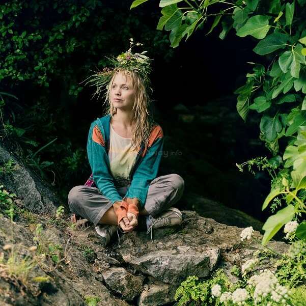 A woman sits cross-legged on a rock by a cave, wearing a crown of wildflowers. She looks peaceful, surrounded by lush greenery and natural beauty.