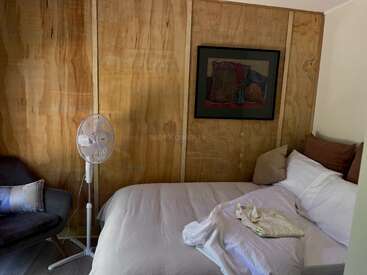 Un acogedor dormitorio con una cama de matrimonio, ropa de cama blanca, almohadas marrones, un ventilador de pie, un sillón oscuro, paredes revestidas de paneles de madera y cuadros enmarcados sobre la cama.