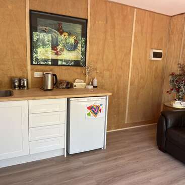 Esta acogedora cocina americana cuenta con armarios blancos, una mininevera decorada con corazones de colores, paredes de madera, una encimera con un hervidor y un cuadro que representa un bodegón con frutas.