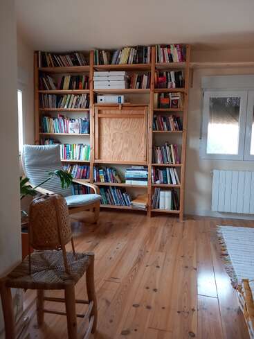 Um canto de leitura aconchegante com estantes de madeira cheias de livros, uma cadeira almofadada, uma cadeira de tecido, piso de madeira, planta de casa, janela e um radiador branco visível.