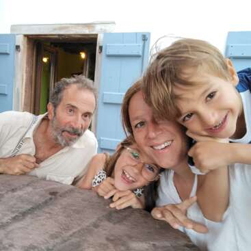 Uma família feliz de quatro pessoas posa junta, sorrindo para a câmera. Eles estão do lado de fora de um prédio com persianas azuis, exalando calor, alegria e união.