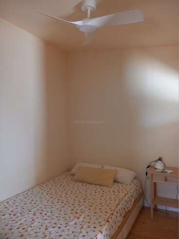 Um quarto pequeno e aconchegante com uma cama de solteiro coberta com uma colcha floral, dois travesseiros, uma mesa de cabeceira, abajur e um ventilador de teto branco moderno.
