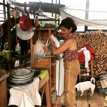 Une femme vêtue d'un haut à fleurs et d'un pantalon beige sèche la vaisselle dans une cuisine rustique, entourée d'assiettes empilées, d'ustensiles de cuisine, de bois de chauffage et d'un petit chien blanc.