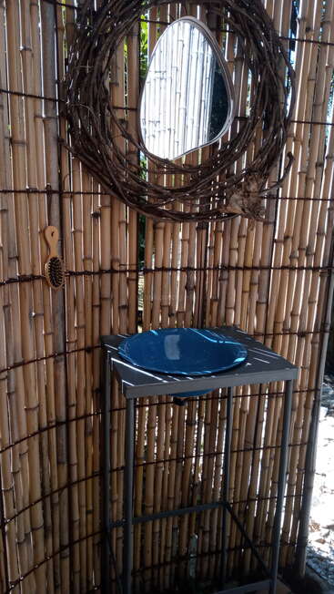 Cette image montre un lavabo extérieur aux murs de bambou avec un bassin bleu, un miroir circulaire encadré de vigne, une brosse à cheveux en bois et la lumière du soleil qui projette des ombres distinctes. Décor rustique et naturel.