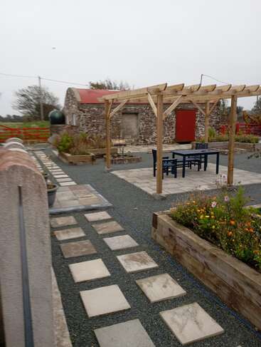 The image shows a neat garden with stone paths, raised flower beds, and a wooden pergola. There’s outdoor seating and a rustic stone building nearby.