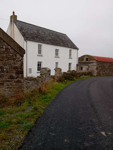 A curved rural road passes by an old white house with a slate roof and stone walls, surrounded by grass and another rustic stone building.