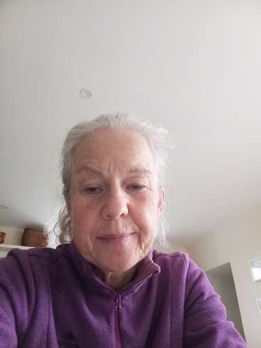 An older woman with gray hair is wearing a purple fleece jacket, sitting indoors. She appears relaxed, looking into the camera, with a neutral facial expression.