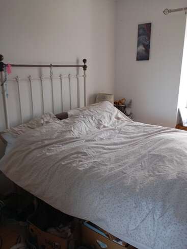 This image shows a bedroom with an unmade bed, white bedding, a metal headboard, some boxes underneath, a small nightstand, and artwork on the wall.