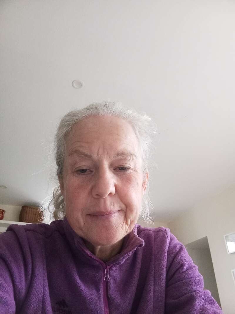 An older woman with gray hair is wearing a purple fleece jacket, sitting indoors. She appears relaxed, looking into the camera, with a neutral facial expression.