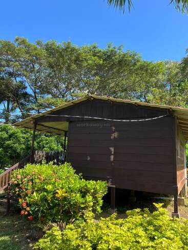 Eine kleine Holzhütte steht auf Stelzen, umgeben von üppigem Grün und blühenden Sträuchern. Hohe Bäume und ein strahlend blauer Himmel vervollständigen die friedliche, tropische Umgebung.