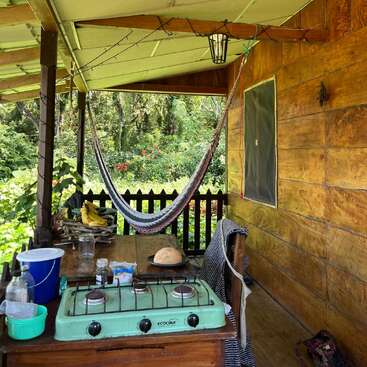 Eine gemütliche Holzveranda mit einer Hängematte, einer einfachen Außenküche mit einem grünen Herd, Brot, Bananen und einem Blick auf den üppigen grünen Garten erinnert an ein friedliches, rustikales Leben.