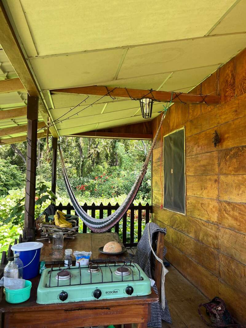 A cozy wooden porch features a hammock, simple outdoor kitchen with a green stove, bread, bananas, and lush green garden view, evoking peaceful rustic living.