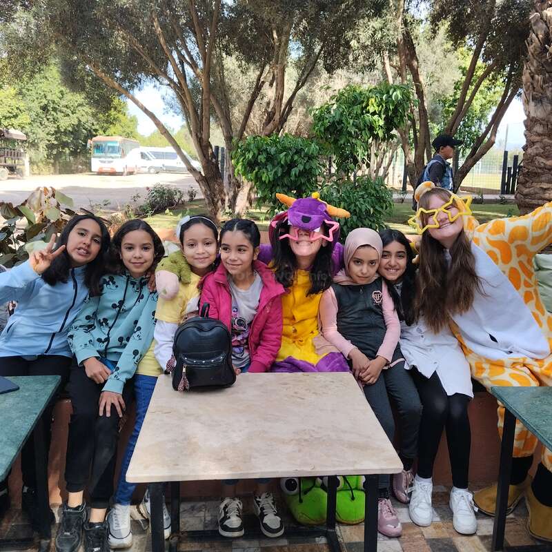 The image depicts a group of seven young girls posing for a photo, with one girl wearing a giraffe costume and another wearing a purple dragon mask.