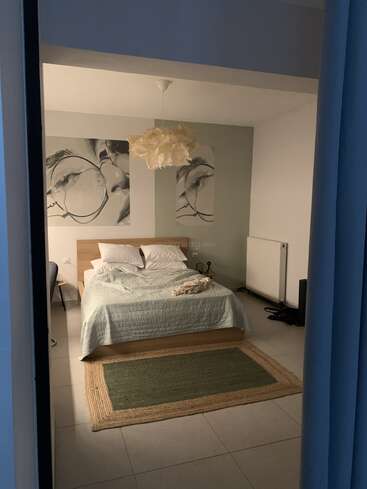 Um quarto aconchegante com uma cama de casal, roupa de cama clara, dois travesseiros, um tapete verde, arte facial abstrata, luminária pendente, radiador, cadeira e piso de ladrilho.
