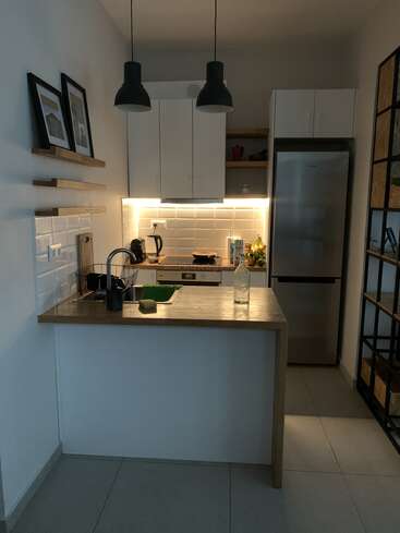 Uma cozinha moderna e compacta com armários brancos, bancadas de madeira, duas lâmpadas pretas penduradas, quadros emoldurados, geladeira de aço inoxidável, prateleiras abertas e iluminação sutil sob os armários.