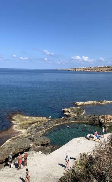 As pessoas aproveitam um dia ensolarado em uma piscina de pedra natural próxima ao mar, com água azul clara, uma costa rochosa e prédios distantes visíveis.
