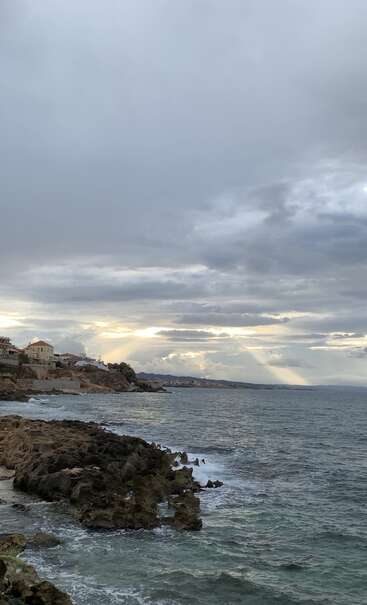 Ein wolkenverhangener Himmel erstreckt sich über einer felsigen Küste, wo sanfte Wellen auf schroffe Ufer treffen. Entfernte Häuser thronen auf den Klippen, das Sonnenlicht bricht durch die Wolken und schafft eine friedliche, dramatische Meereslandschaft.