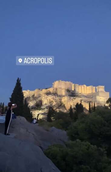 Die beleuchtete Akropolis thront in der Abenddämmerung majestätisch auf einem felsigen Hügel, umgeben von Bäumen und Besuchern, die diesen historischen Anblick unter einem klaren Abendhimmel festhalten.