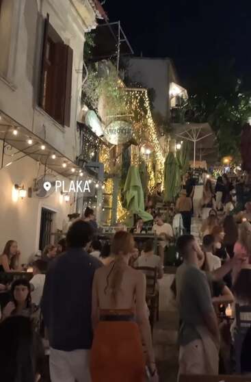 Das Bild zeigt eine lebhafte Nachtszene in Plaka, Athen, mit Menschen, die im Freien essen, Lichterketten, einer Treppe und einer pulsierenden, festlichen Atmosphäre unter dem Nachthimmel.