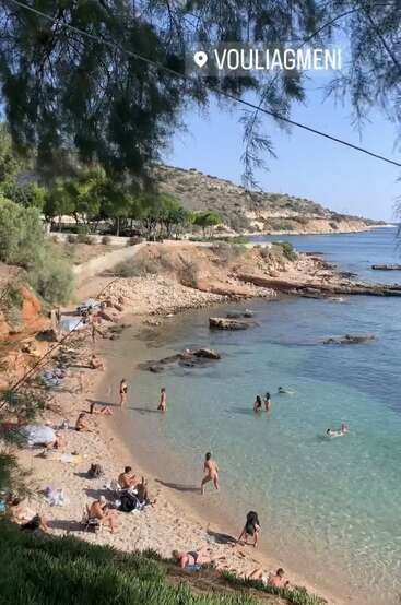 Eine wunderschöne Strandszene in Vouliagmeni, Griechenland. Die Menschen entspannen sich am Sandstrand und schwimmen im klaren, blauen Wasser, umgeben von Bäumen und felsigen Hügeln.