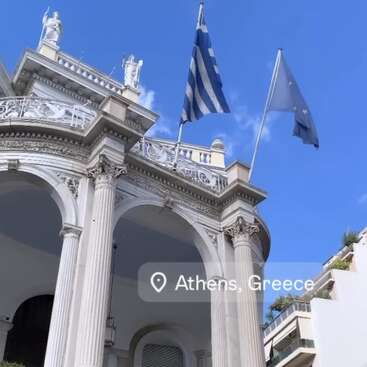 This image shows an elegant neoclassical building in Athens, Greece, with Greek and EU flags, ornate columns, statues, and a bright blue sky above.