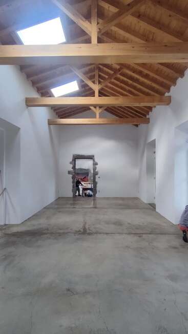Esta imagen muestra una habitación espaciosa con paredes blancas, suelo de hormigón pulido, techo de vigas de madera, tragaluces y una puerta con marco de piedra y una persona dentro.