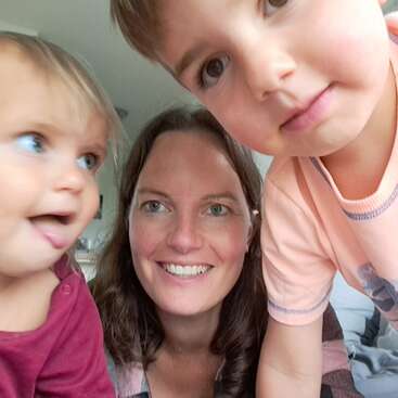 Una mujer sonriente posa para un selfie con dos niños pequeños. Uno de los niños parece juguetón y enérgico, mientras que el otro mira con curiosidad a la cámara. Una escena alegre y luminosa.