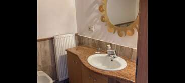 Cette salle de bains comprend un comptoir en marbre avec un lavabo blanc, des armoires en bois, un miroir rond décoratif, un distributeur de savon, un radiateur et une vue partielle des toilettes.