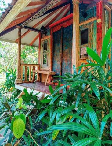 A rustic wooden cabin with colorful, weathered walls and intricate carvings sits surrounded by lush green tropical plants, exuding charm and natural tranquility.