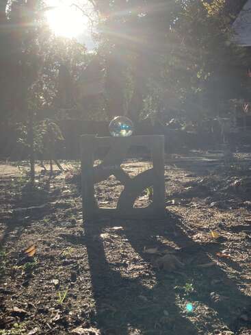 Sunlight streams through trees, casting long shadows over a concrete block with artistic cutouts. A reflective glass sphere rests atop, creating a magical, serene atmosphere.