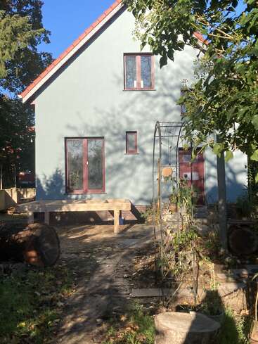 A two-story house with light blue walls and red-framed windows, surrounded by trees and a garden. Wooden bench, log, and plant trellis in front.