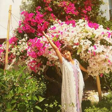 The image depicts a woman standing in front of a vibrant bush with pink and white flowers, her arms raised in a joyful gesture.