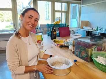 A smiling woman stands in a bright kitchen, preparing dough in a round baking tin. Various kitchen items, food, and utensils are scattered across the wooden counter.