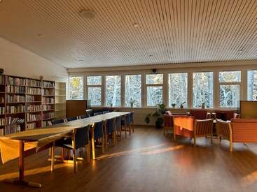 A cozy room with wooden ceiling, bookshelves, long table, blue and orange chairs, large windows, houseplants, and snowy trees outside bathed in warm sunlight.