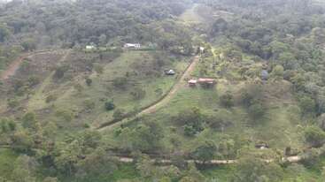 Vista aérea de un paisaje exuberante, verde y montañoso, con casas dispersas, caminos de tierra y un denso bosque que rodea campos abiertos y parcelas de tierra cultivada. Paisaje tranquilo.
