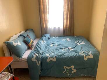 Um quarto pequeno e aconchegante com paredes amarelas apresenta uma cama bem arrumada, coberta com roupas de cama azuis com estampa de estrelas, posicionada ao lado de uma janela com cortinas marrons claras.