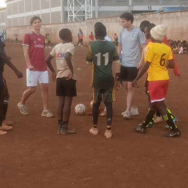 Um grupo de jovens, alguns com uniformes esportivos, se reúne em círculo em um campo de futebol de terra, preparando-se para um jogo ou sessão de prática.