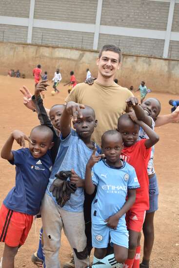 Eine Gruppe lächelnder Kinder und ein Erwachsener posieren zusammen auf einem schmutzigen Fußballfeld. Andere Kinder spielen im Hintergrund und zeigen eine fröhliche, freundliche Atmosphäre.