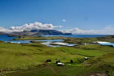 Rolling green hills dotted with small buildings lead to a serene, blue coastal inlet. Distant mountains and dramatic clouds complete this peaceful, scenic landscape view.