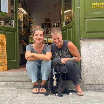 The image depicts two women and a dog sitting outside a green storefront, with the woman on the left wearing a grey shirt and jeans, and the other in a Star Wars tank top and black pants.