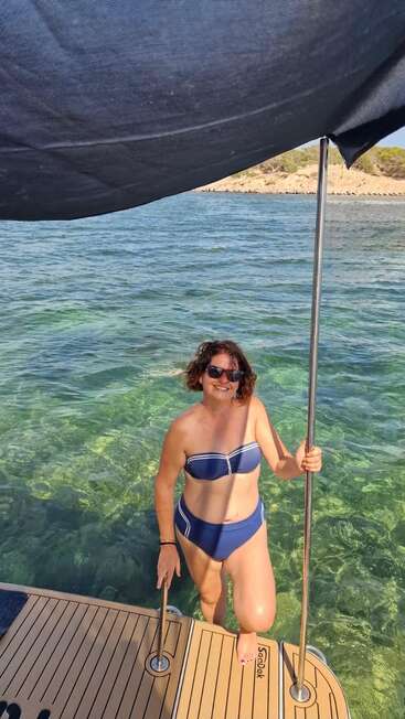 A woman in a blue bikini steps onto a boat from clear turquoise water, smiling, wearing sunglasses, under a large black sunshade, enjoying a sunny day.