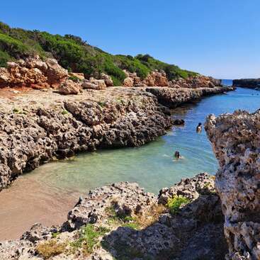 A beautiful rocky coastline with clear turquoise water, lush green shrubs atop the cliffs, a small sandy cove, and a few people swimming under a bright blue sky.