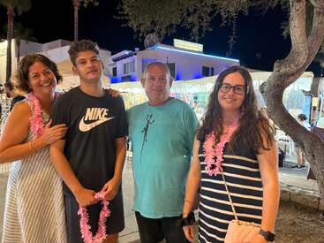 Four people, smiling and standing together at night by market stalls. They wear leis, enjoying a festive outdoor atmosphere with bright lights and a relaxed mood.