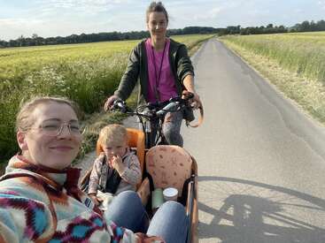 Três pessoas, incluindo uma criança, andam em uma bicicleta de carga em uma pacífica estrada rural. Os adultos estão sorrindo, curtindo o dia ensolarado e os campos panorâmicos.